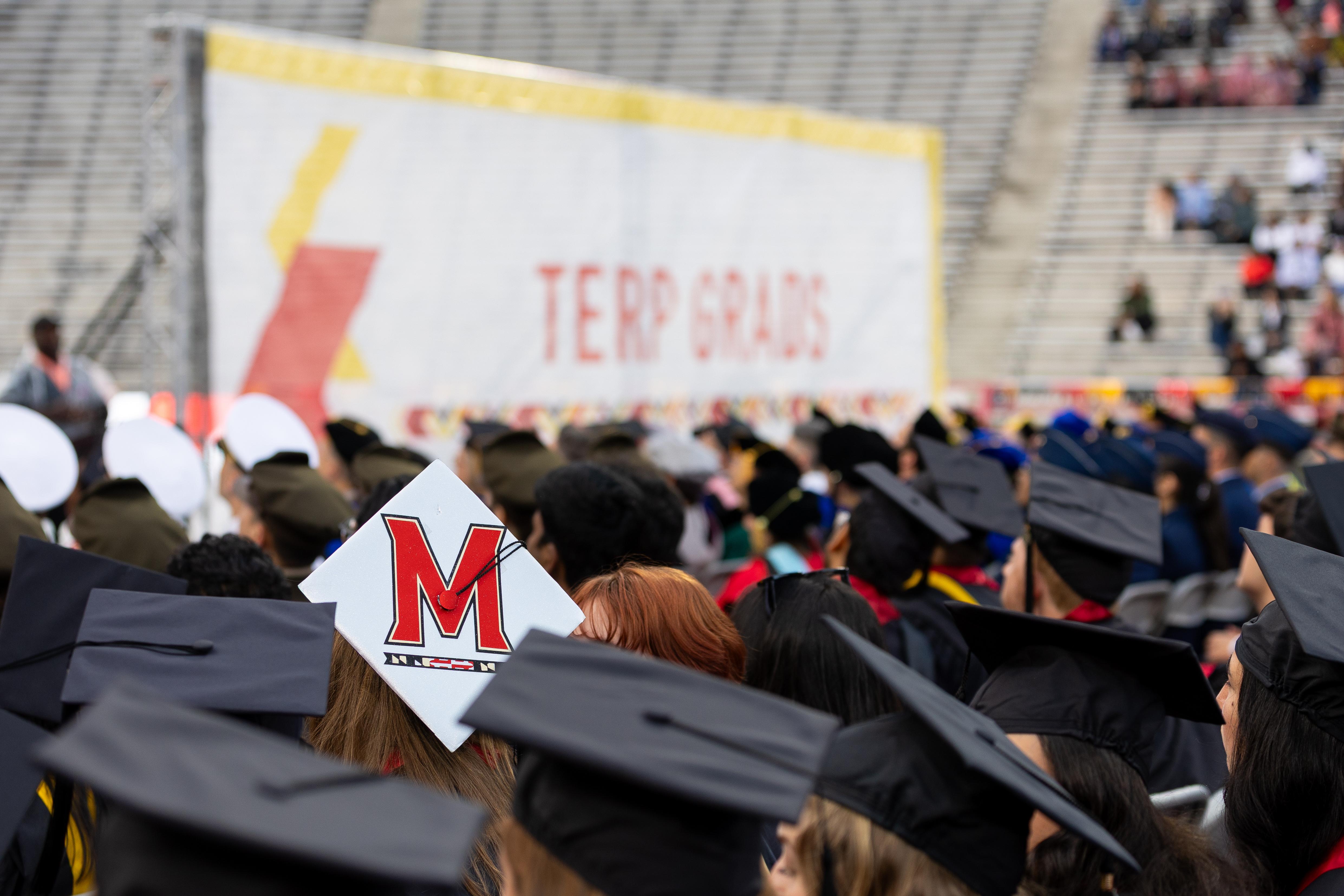 UMD Commencement image from 2025. Many graduates with one grad cap with a red "M" facing Terp Grads sign.